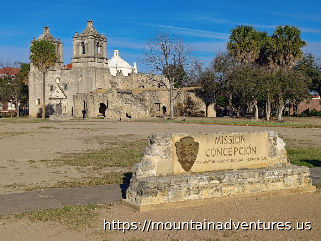 The NPS Entrance sign for Mission Conception Area of San Antonio Missions with the Church behind it.